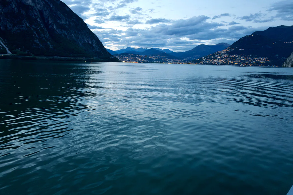 Lake Lugano and its mountain shoreline as evening lights begin to appear