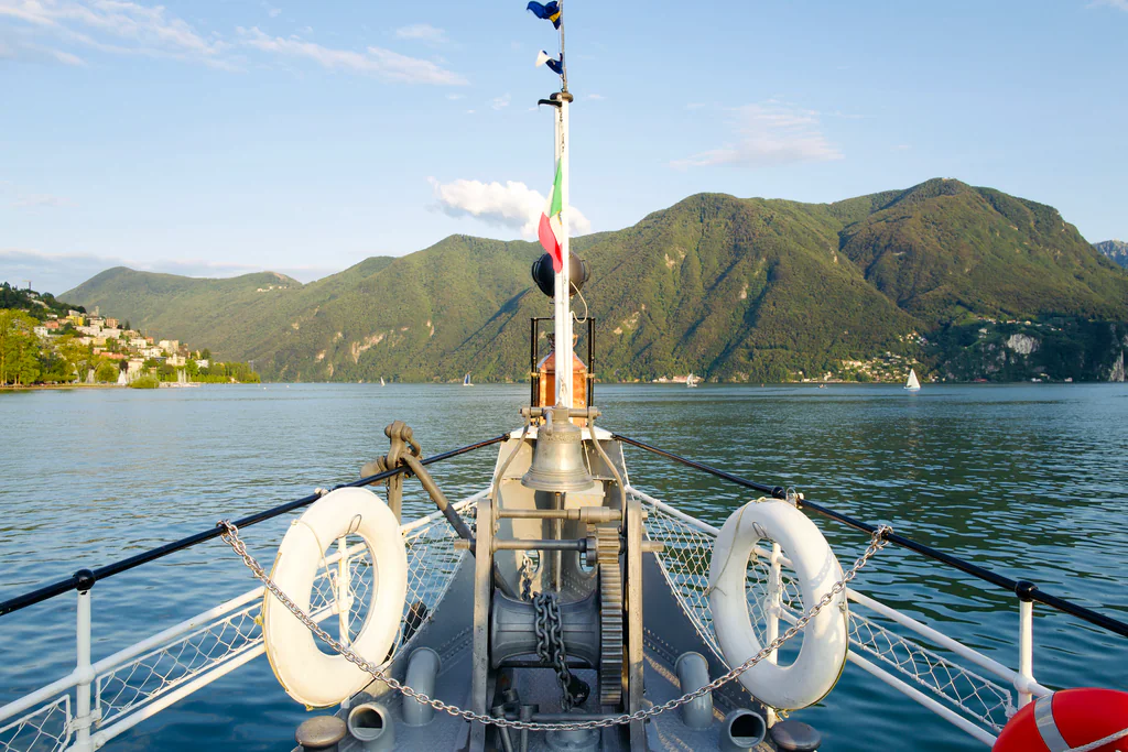 The bow of a lake steamboat crossing Lake Lugano toward the mountains